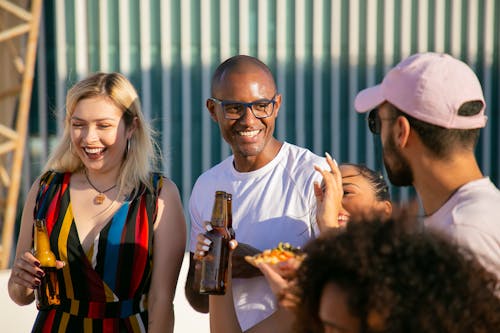 Group of delighted young multiracial friends in summer clothes drinking cold beer and chatting while chilling together on sunny backyard