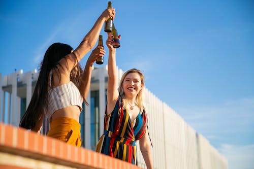Delighted young female friends in stylish summer outfits toasting with beer bottles and smiling while spending sunny weekend on rooftop
