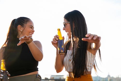 Attractive joyful female friends in summer wear enjoying cold beer and chatting happily while looking at each other with laughter