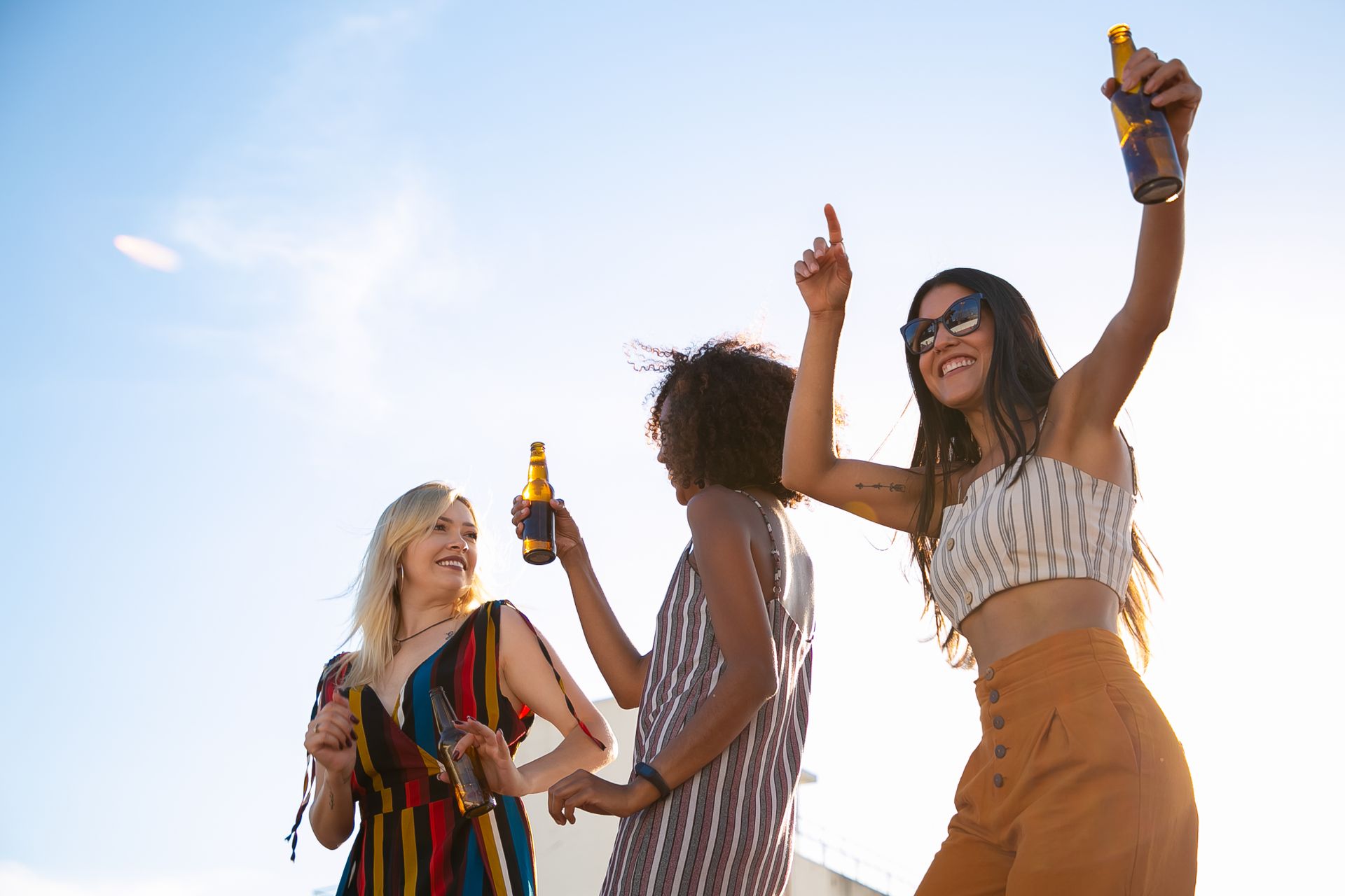 Three diverse young women celebrating with drinks on a sunny rooftop party.