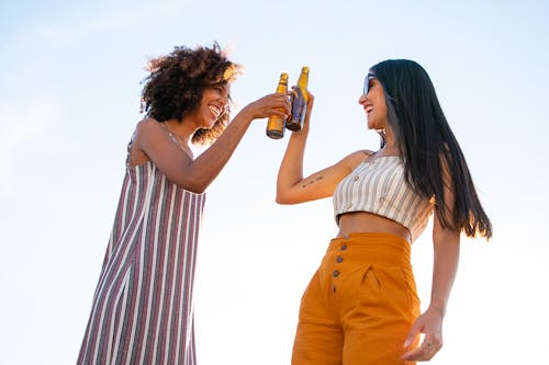 Low angle of delighted multiracial female friends toasting with bottles of beer while laughing together during party