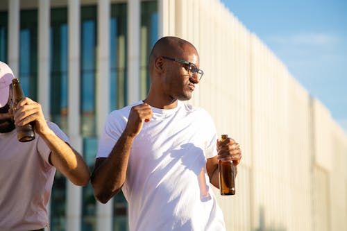 Positive young multiracial male friends drinking bottles of beer and dancing during open air party on sunny day