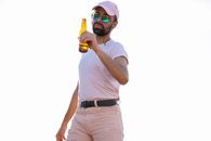 Masculine ethnic male in trendy wear and modern sunglasses holding bottle of alcoholic drink while looking at camera on white background