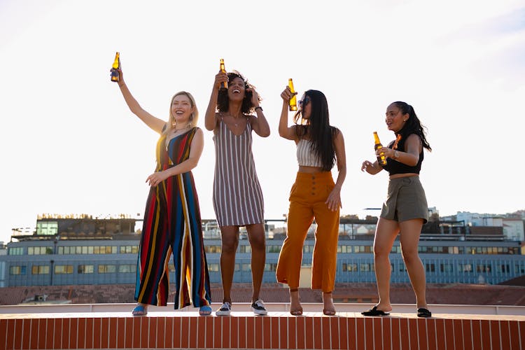 Cheerful Young Women Drinking Beer And Dancing On Rooftop