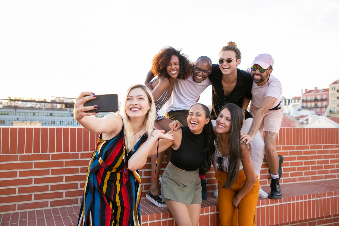 Free Group of cheerful young male and female multiracial friends laughing and taking selfie on smartphone while spending time together on terrace Stock Photo