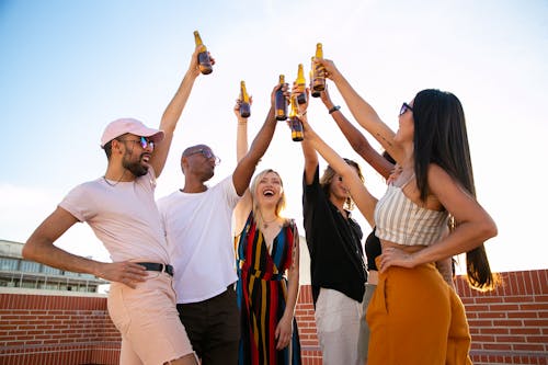 Group of cheerful multiethnic friends clinking beer bottles during event on terrace