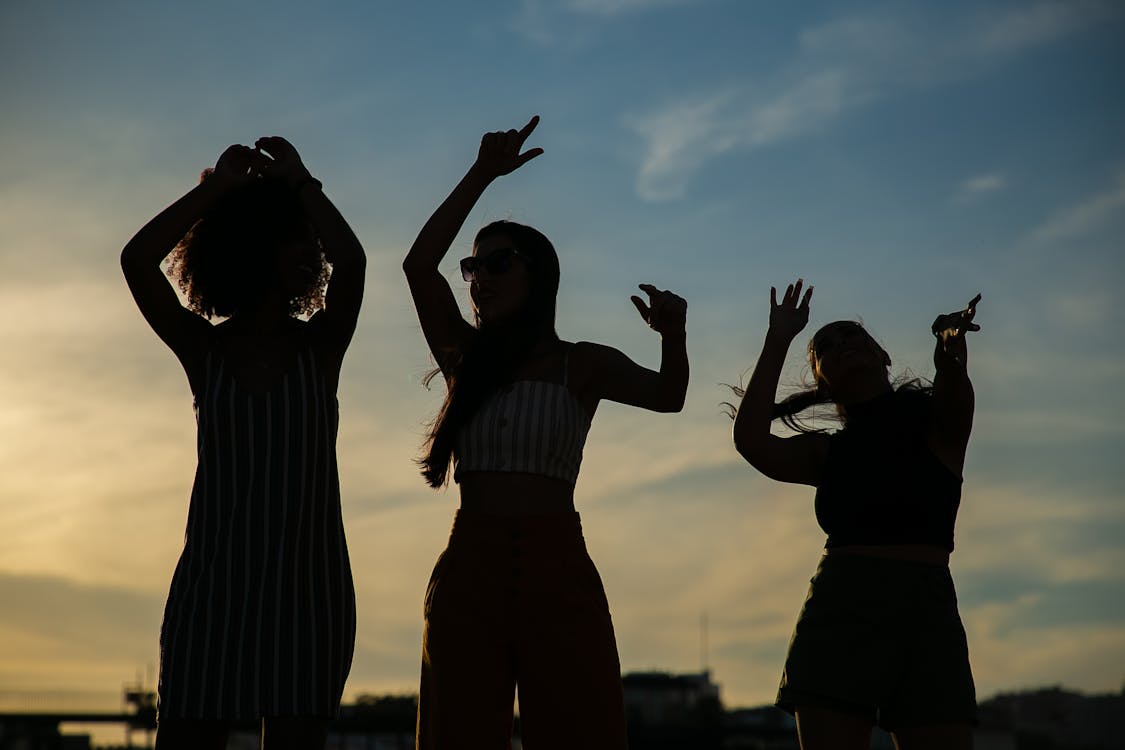 Free Low angle silhouettes of unrecognizable young female friends dancing against cloudy sunset sky during open air party Stock Photo