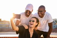 Cheerful young multiethnic men laughing and taking selfie on smartphone in sunlight