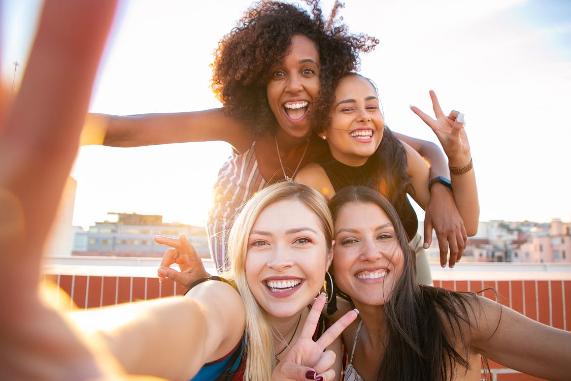 Free Cheerful young diverse women showing V sign while taking selfie on rooftop Stock Photo