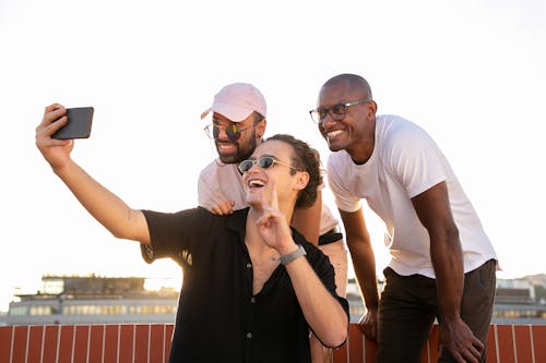 Low angle of positive young guy showing peace sign and smiling while taking selfie on smartphone with diverse friends on building rooftop