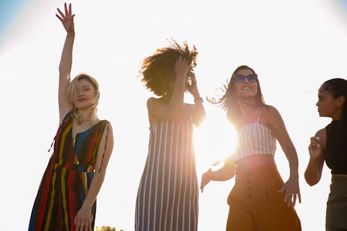 From below of young content diverse ladies in trendy outfits smiling and dancing against cloudless sky during open air party on sunny day