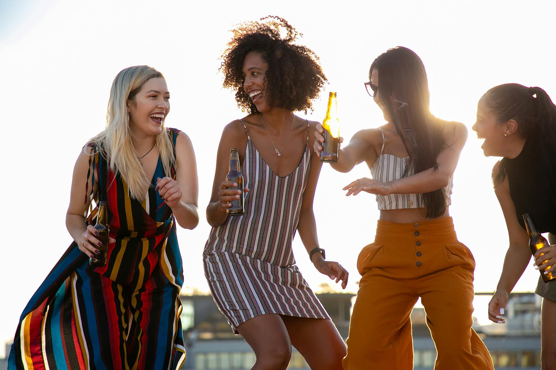 Group of cheerful young multiracial ladies laughing and dancing with beer bottles in hands during summer party on rooftop on sunny day