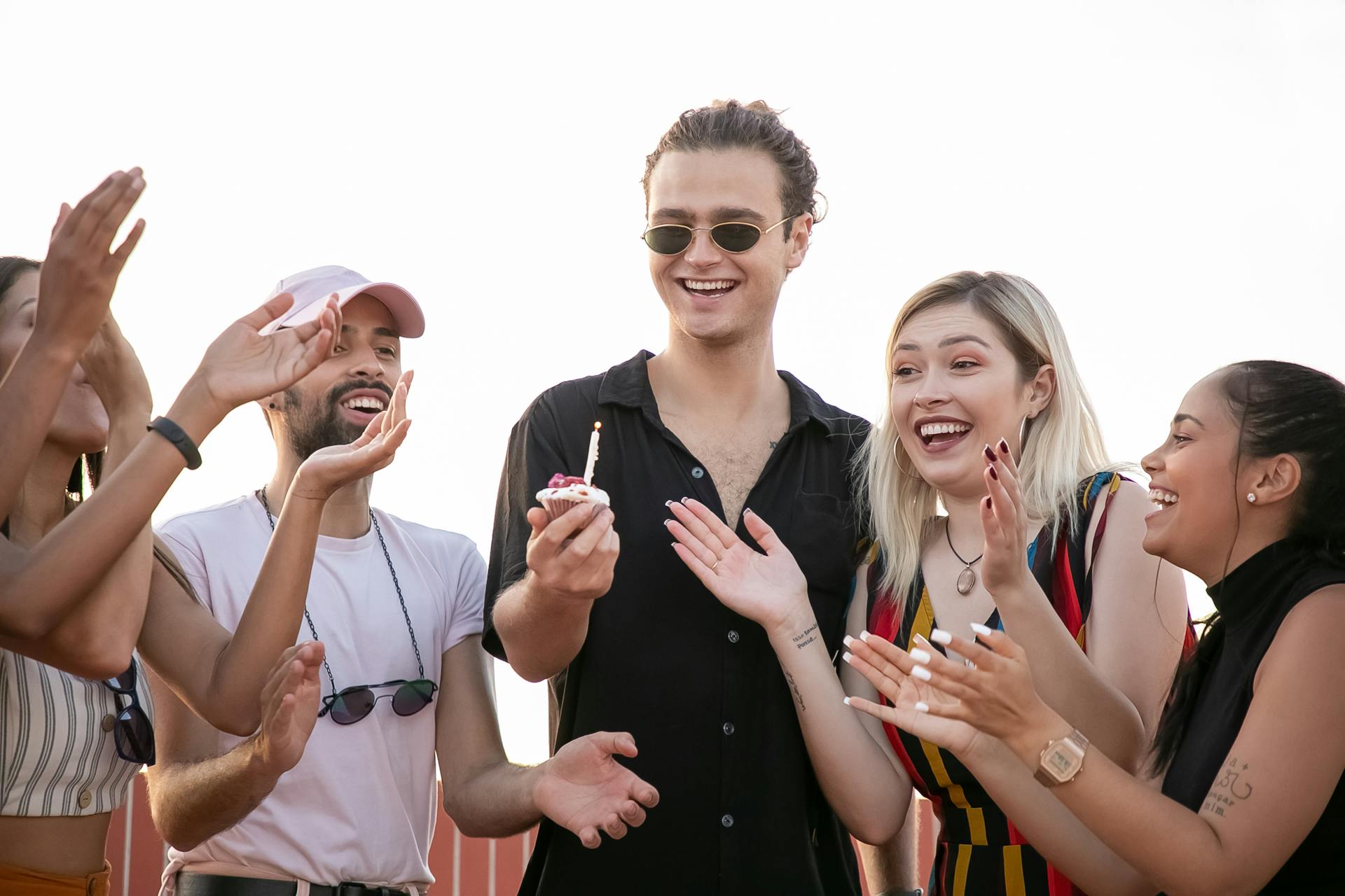 Happy young stylish guy in shirt and sunglasses holding cupcake with candle in hand while making wish during birthday party with cheerful diverse friends