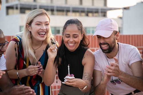 Cheerful young diverse friends laughing and clapping hands while young ethnic lady making wish and blowing out candle on cupcake during open air birthday party