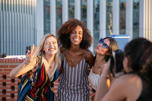 Unrecognizable female photographer taking picture of cheerful young multiracial female friends smiling and showing V sign during party on modern building rooftop