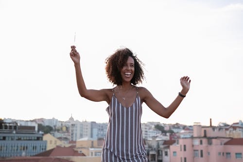 Cheerful young African American female with curly hair in stylish dress dancing with sparkler in had during open air party on rooftop in modern city