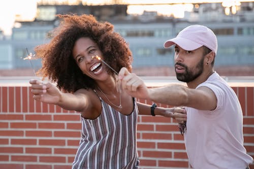 Joyful young African American female and ethnic guy having fun while dancing with sparklers in hands during open air party on modern building terrace