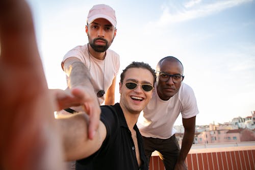 Cheerful young guy in casual outfit and sunglasses taking selfie with multiracial male friends while spending time together on modern building rooftop
