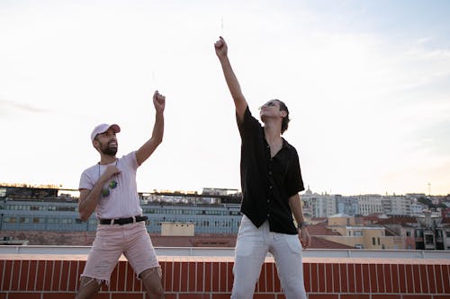 Positive young multiethnic male friends in stylish outfits dancing on rooftop with sparklers in hands during party on building rooftop