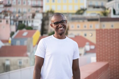 Free Positive young hairless African American male in white t shirt and eyeglasses smiling while standing on building rooftop on sunny day Stock Photo