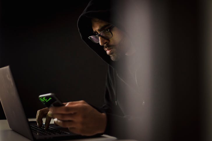 Crop ethnic hacker with smartphone typing on laptop in darkness