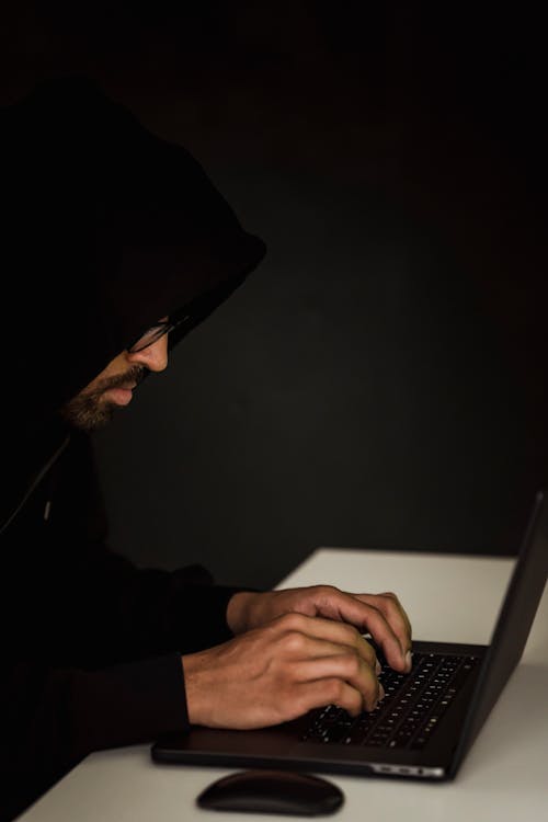 Side view of crop anonymous young ethnic male cyber spy in hoodie typing on portable computer on black background