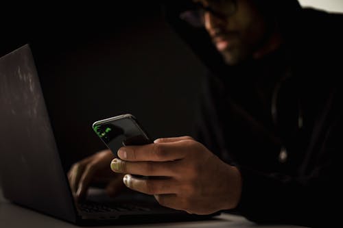 Free Crop anonymous ethnic male cyber spy with cellphone and netbook hacking system in evening Stock Photo