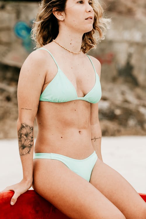 Crop young contemplative female surfer in swimwear sitting on surfboard on beach while looking away