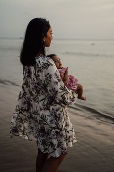 日落时分，一位母亲穿着花卉连衣裙，在宁静的海边抱着她的孩子
