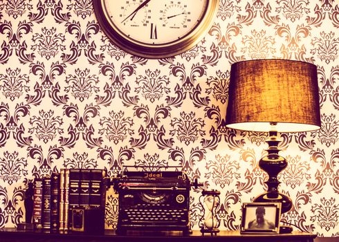 Classic vintage desk setting with typewriter, books, lamp, and clock on ornate wallpaper.