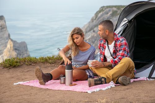 Free A couple enjoys a relaxing picnic with a scenic ocean view, camping on a cliffside with a thermos and tent. Stock Photo