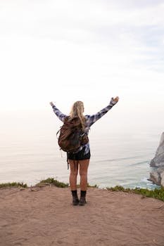 Blonde woman enjoying view atop a cliff in Portugal, arms raised in triumph.