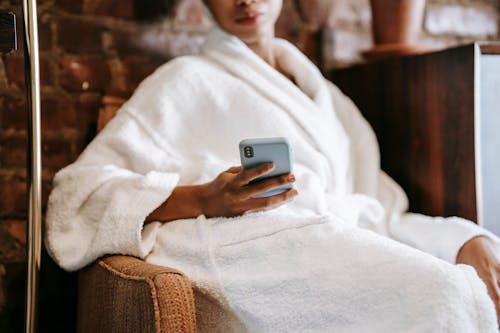 Crop anonymous ethnic female in bathrobe text messaging on cellphone while resting in armchair in beauty salon