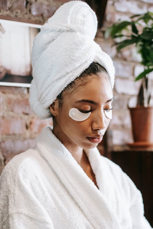 Young ethnic female in terry towel on head and robe resting in spa center during facial procedure