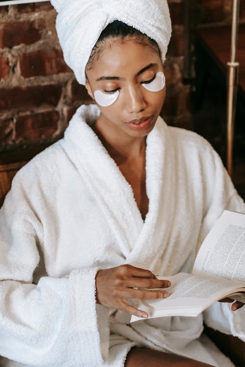 Young focused ethnic female in bathrobe and terry towel with eye patches reading textbook during procedure in spa center