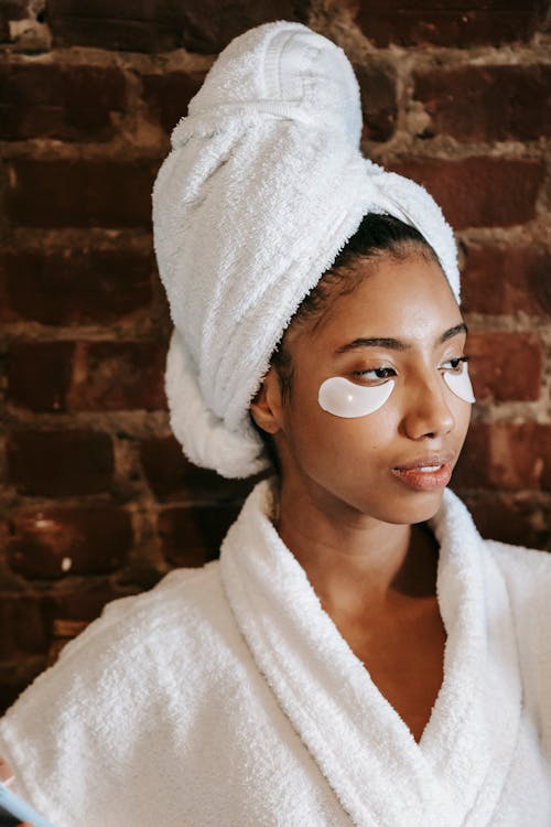 Young contemplative ethnic female in terry towel and robe looking away during facial procedure in beauty salon