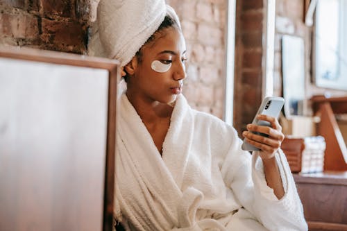 Crop young focused ethic lady in eye patch and terry towel browsing internet on mobile phone during procedure in beauty salon