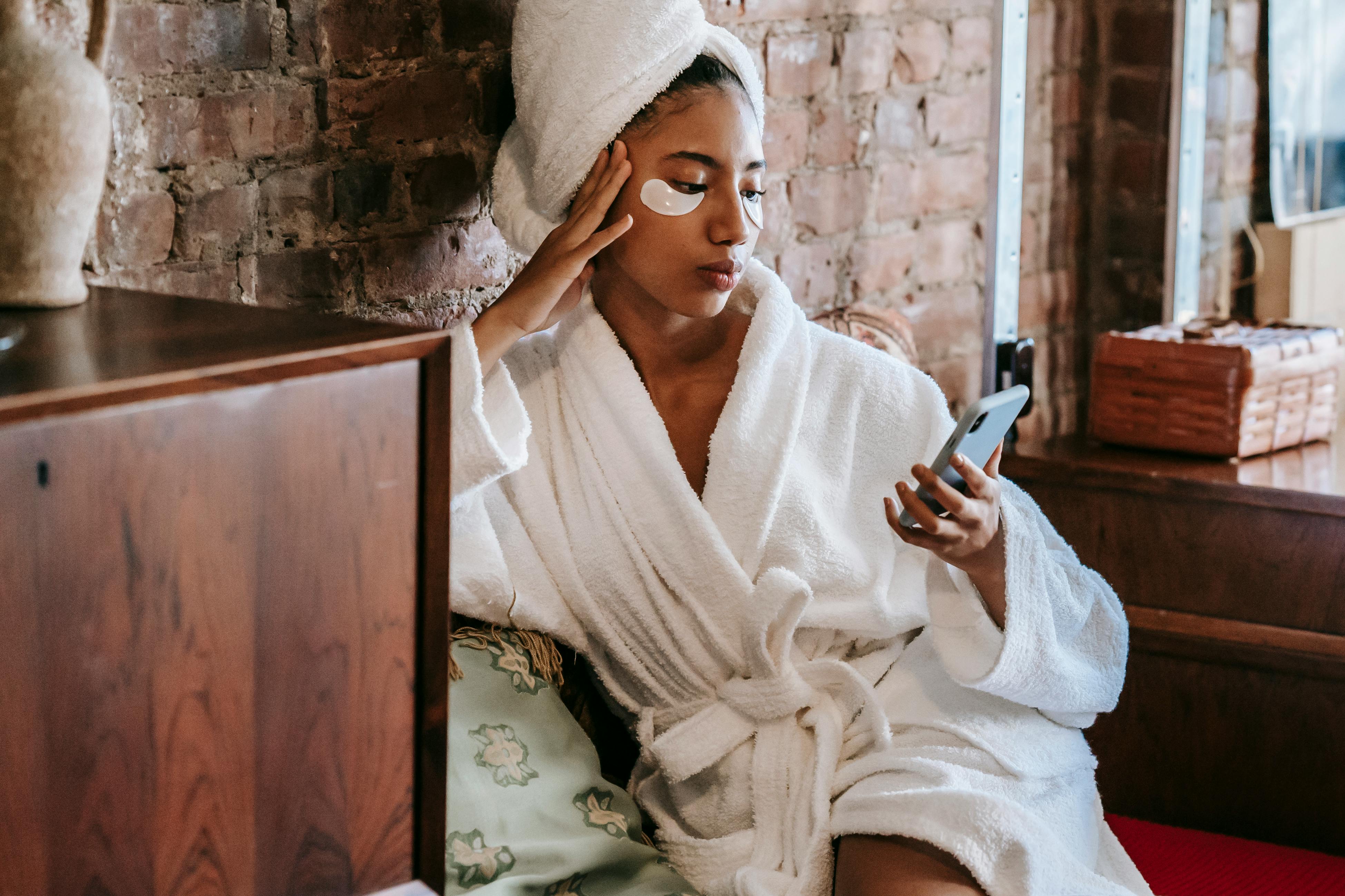 Crop ethnic woman with eye patches and smartphone in salon