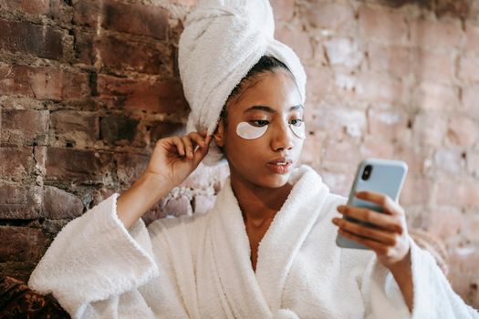 Young attentive ethnic female touching terry towel while surfing internet on cellphone near brick wall in beauty salon