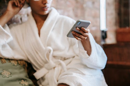 Free Crop anonymous ethnic female in robe leaning on hand while browsing internet on cellphone during facial procedure in spa center Stock Photo