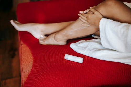 Free High angle of crop unrecognizable barefoot female applying moisturizing cream on leg while sitting on soft sofa Stock Photo