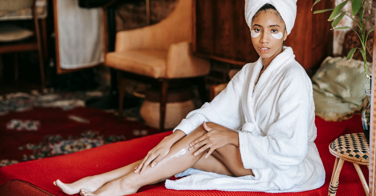 Young barefoot ethnic female in towel and robe applying lotion on leg while sitting on sofa and looking at camera