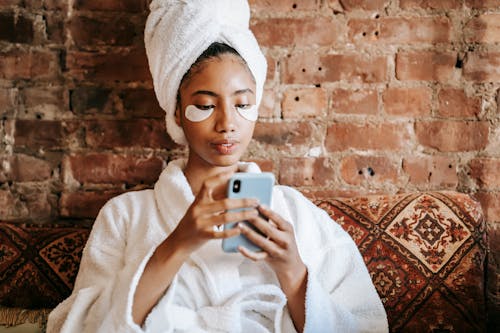 Ethnic female in robe and towel surfing internet on mobile phone during facial procedure near brick wall in house