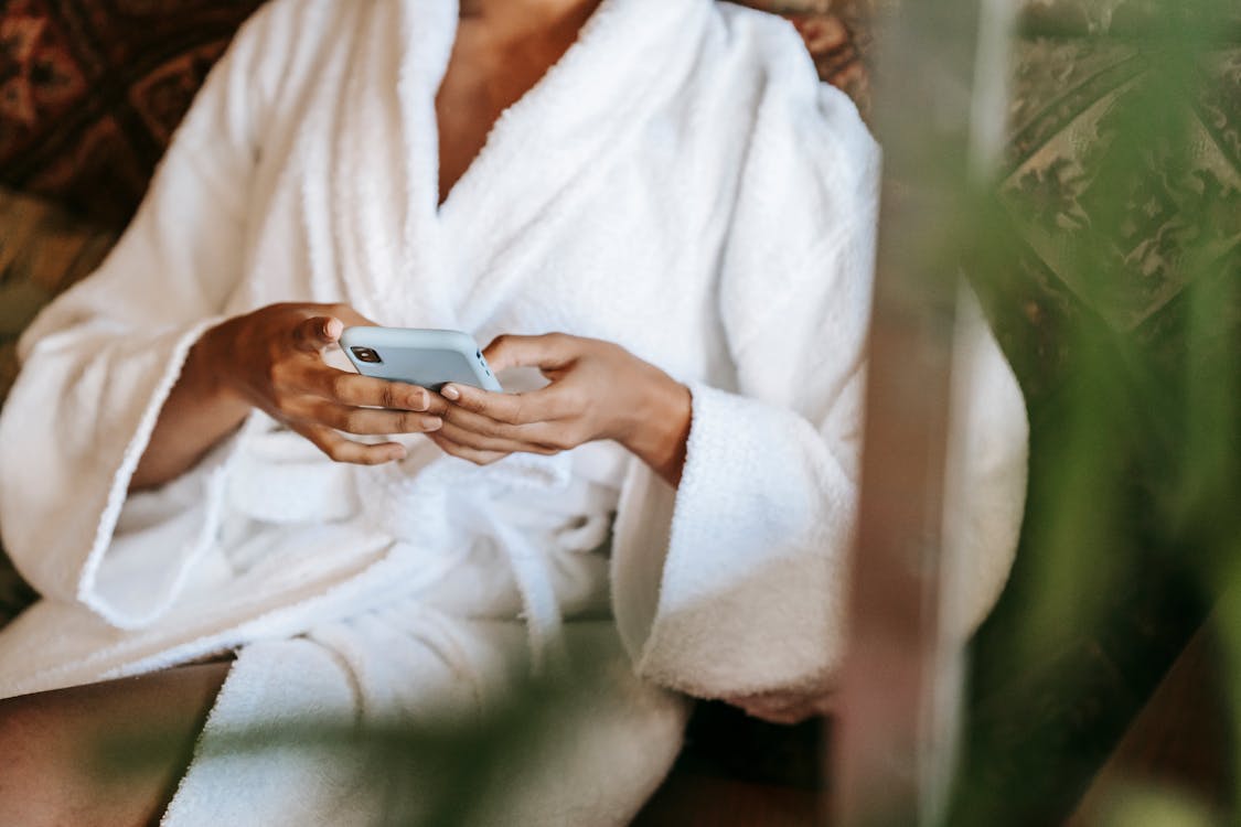 Crop anonymous ethnic female text messaging on cellphone while sitting in beauty salon in sunlight