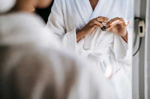 Crop unrecognizable young lady in white bathrobe opening face cream while standing in front of mirror after taking shower in morning