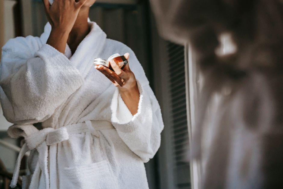 Crop anonymous ethnic female in soft bathrobe applying nourishing cream on face while reflecting in mirror at home