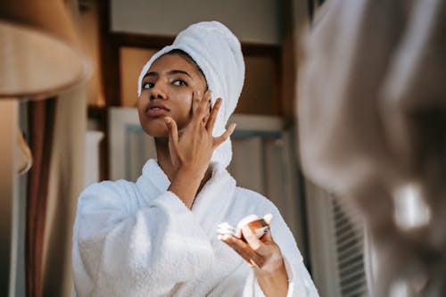 Free Crop young ethnic female in bathrobe with terry towel on head applying smooth cream on face while looking in mirror at home Stock Photo