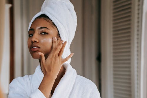 Young ethnic female in robe with terry towel on head applying moisturizing cream on face while looking in mirror in house