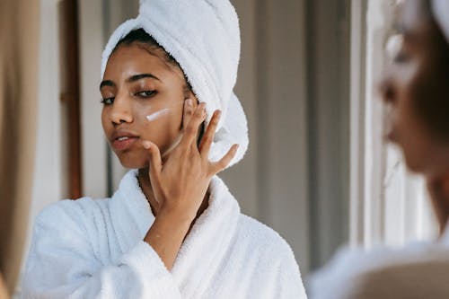 Crop attentive ethnic female in soft robe applying smear of moisturizing cream on cheek against mirror at home