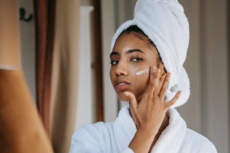 Ethnic Woman Applying Nourishing Cream On Face Against Mirror